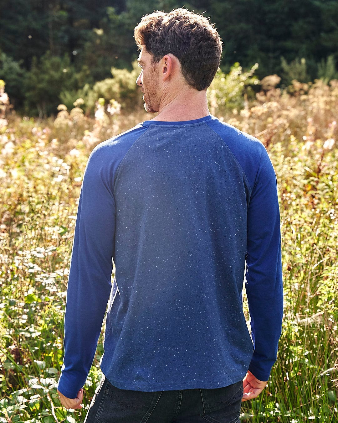 A man standing in a field wearing a blue long sleeved Saltrock Home Run Nep - Men's Raglan Long Sleeve T-Shirt.