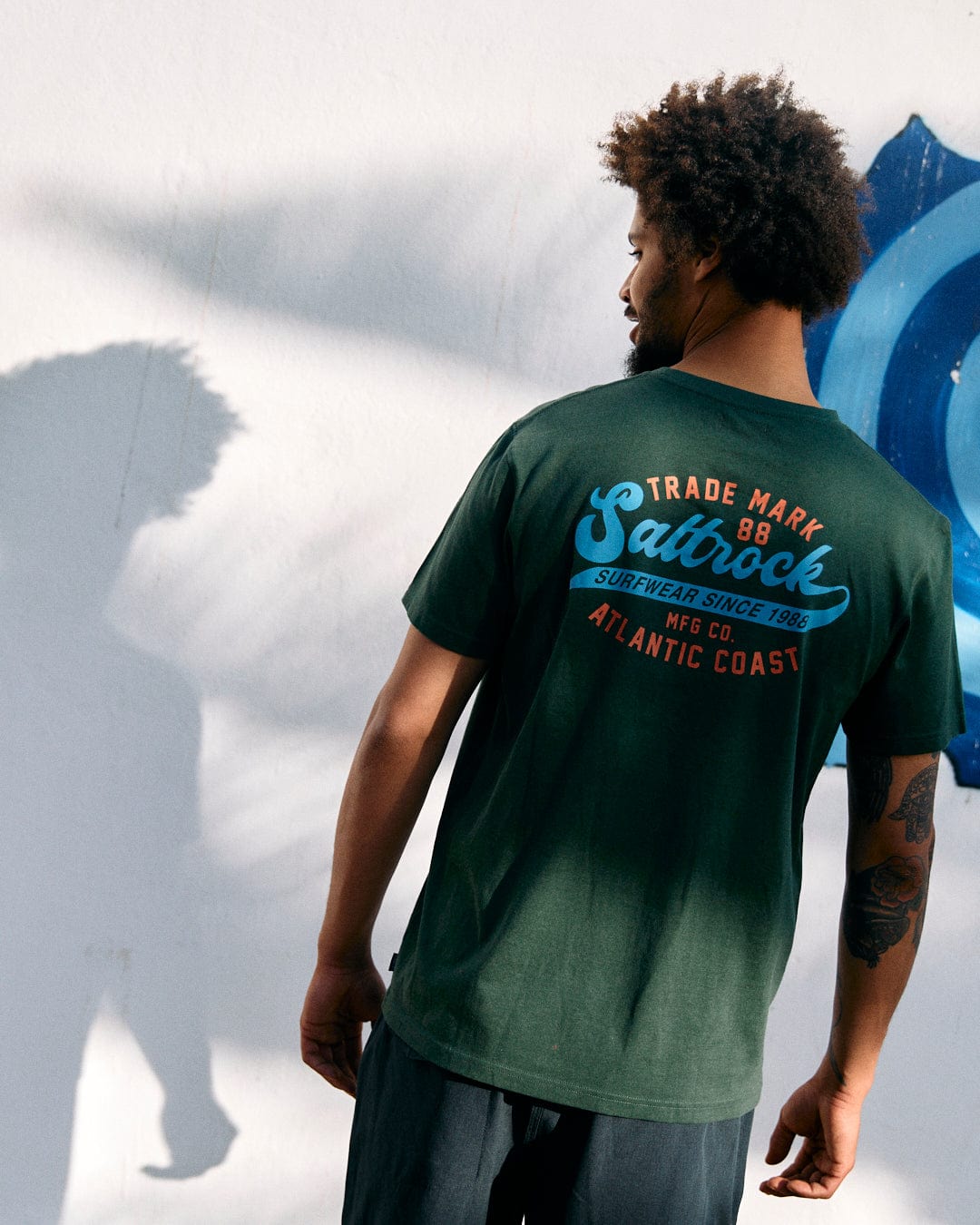 Man viewed from behind, wearing a Saltrock "Home Run" Mens Short Sleeve T-Shirt in Dark Green and casual shorts, standing near a white wall with graffiti. Sun casts his shadow on the wall.