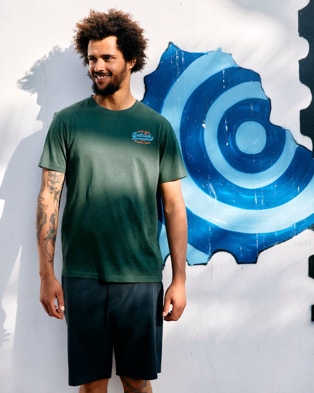 Smiling young man with curly hair, wearing a Saltrock Home Run Mens Short Sleeve T-Shirt in Dark Green and black shorts, standing in front of a wall with a blue abstract mural.