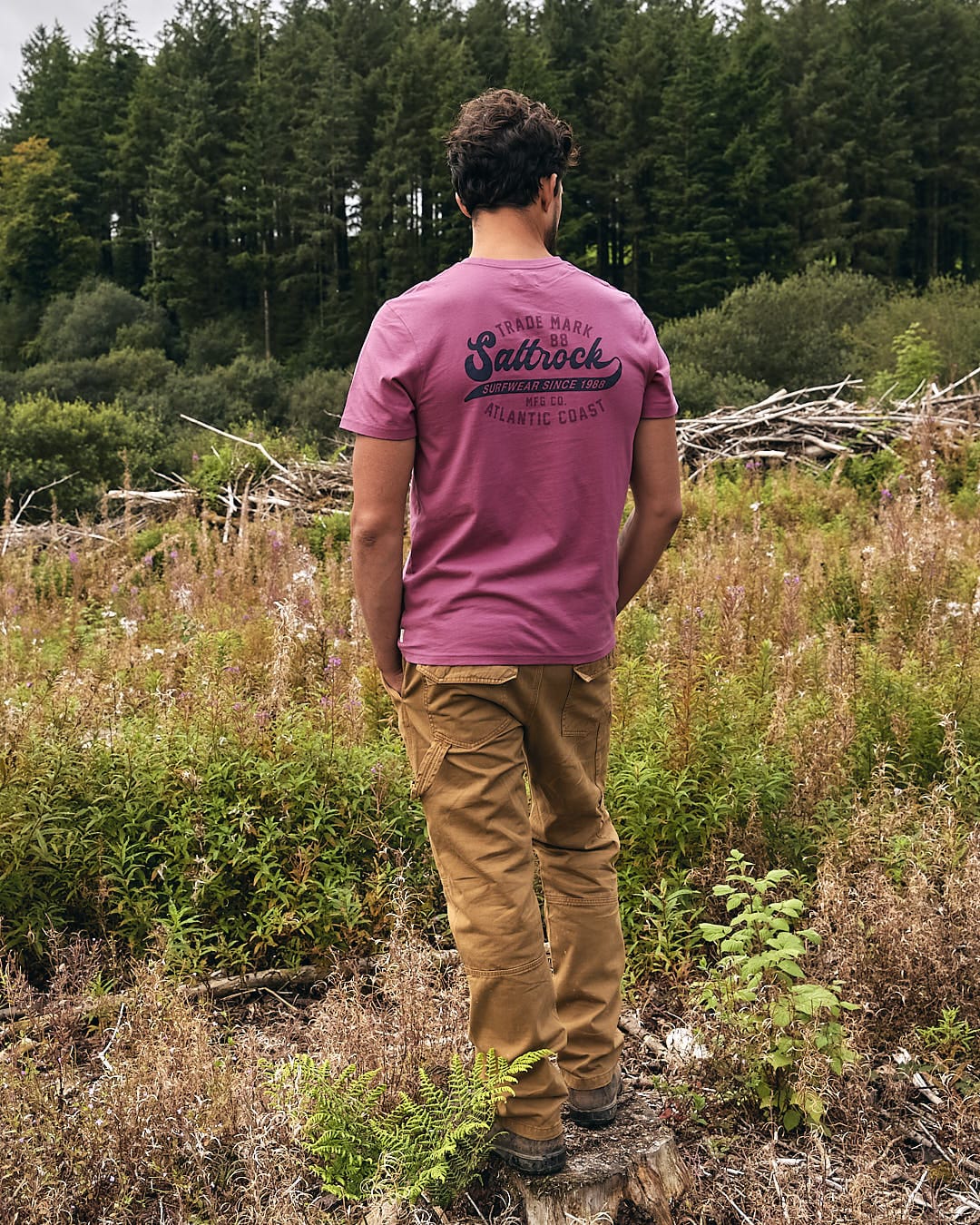 A man wearing a Saltrock Home Run - Mens Short Sleeve T-Shirt - Dark Pink is standing in a wooded area.