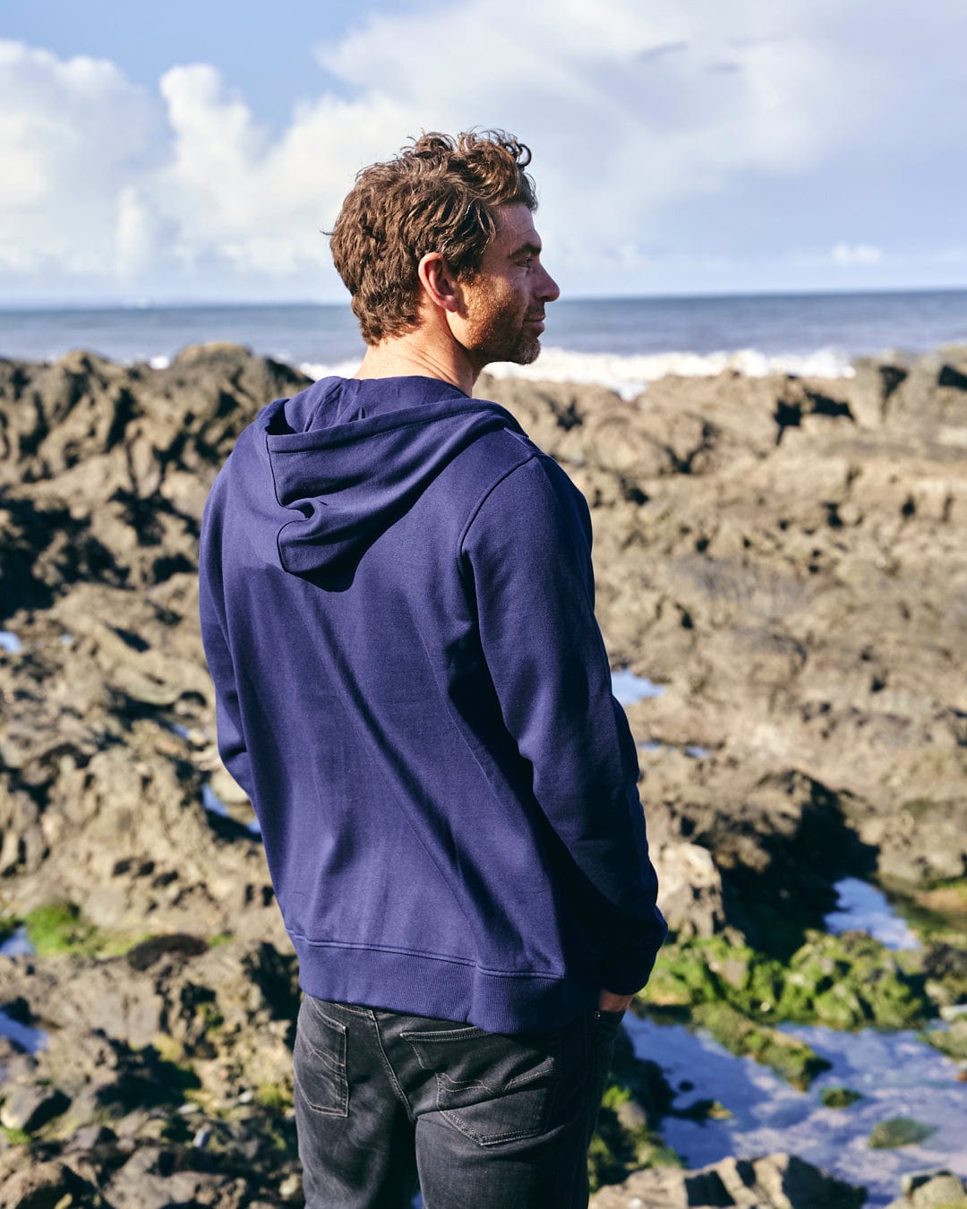 A man standing on rocks looking out to the ocean, wearing a Saltrock Home Run - Mens Zip Hoodie in Blue with drawstring hood.