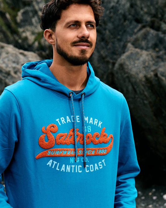 A man wearing the Saltrock Home Run Chenille Men's Pop Hoodie in blue, created from recycled polyester, stands outdoors near rocks and gazes to the side.
