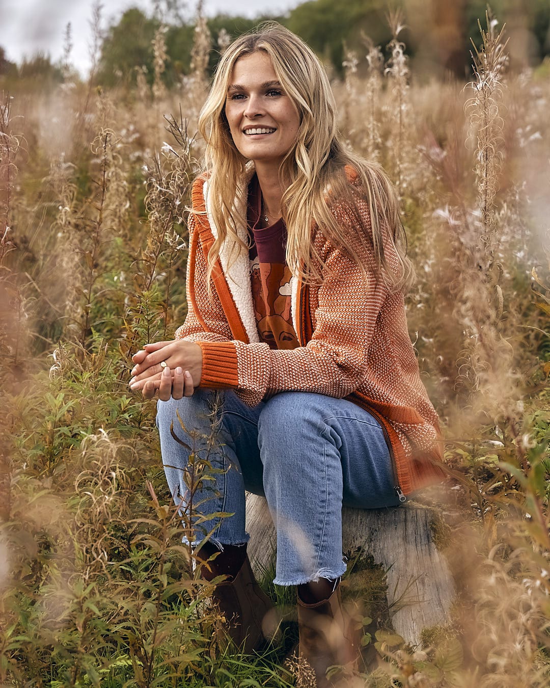 A blonde woman sitting on a log in a field of tall grass wearing the Saltrock - Helly Womens Borg Lined Knitted Hoodie - Orange.
