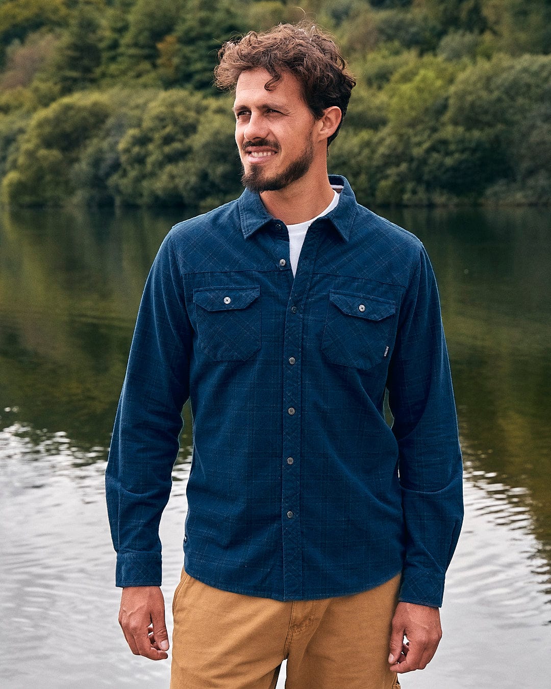 A man standing next to a lake wearing the Saltrock Jaxon - Mens Check Long Sleeve Shirt in Dark Blue.