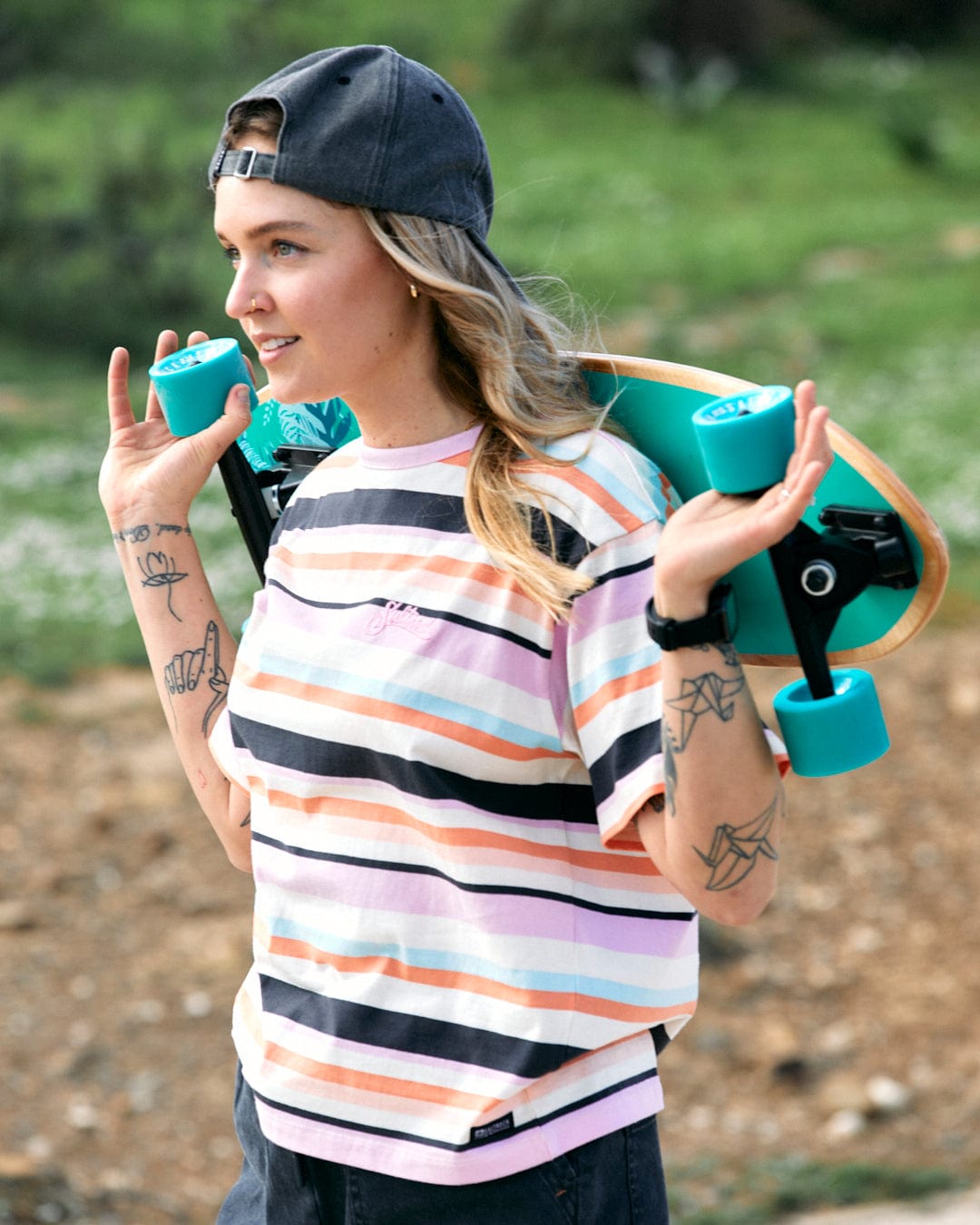A person with a backward baseball cap carrying a skateboard with a Juno - Womens Short Sleeve T-Shirt - Multi design over their shoulder by Saltrock.
