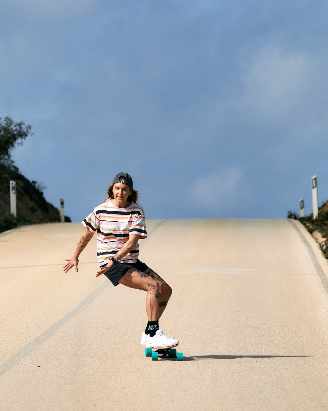 A skateboarder cruising downhill on an open road, his Juno - Womens Short Sleeve T-Shirt - Multi fluttering with a stripe pattern from Saltrock.