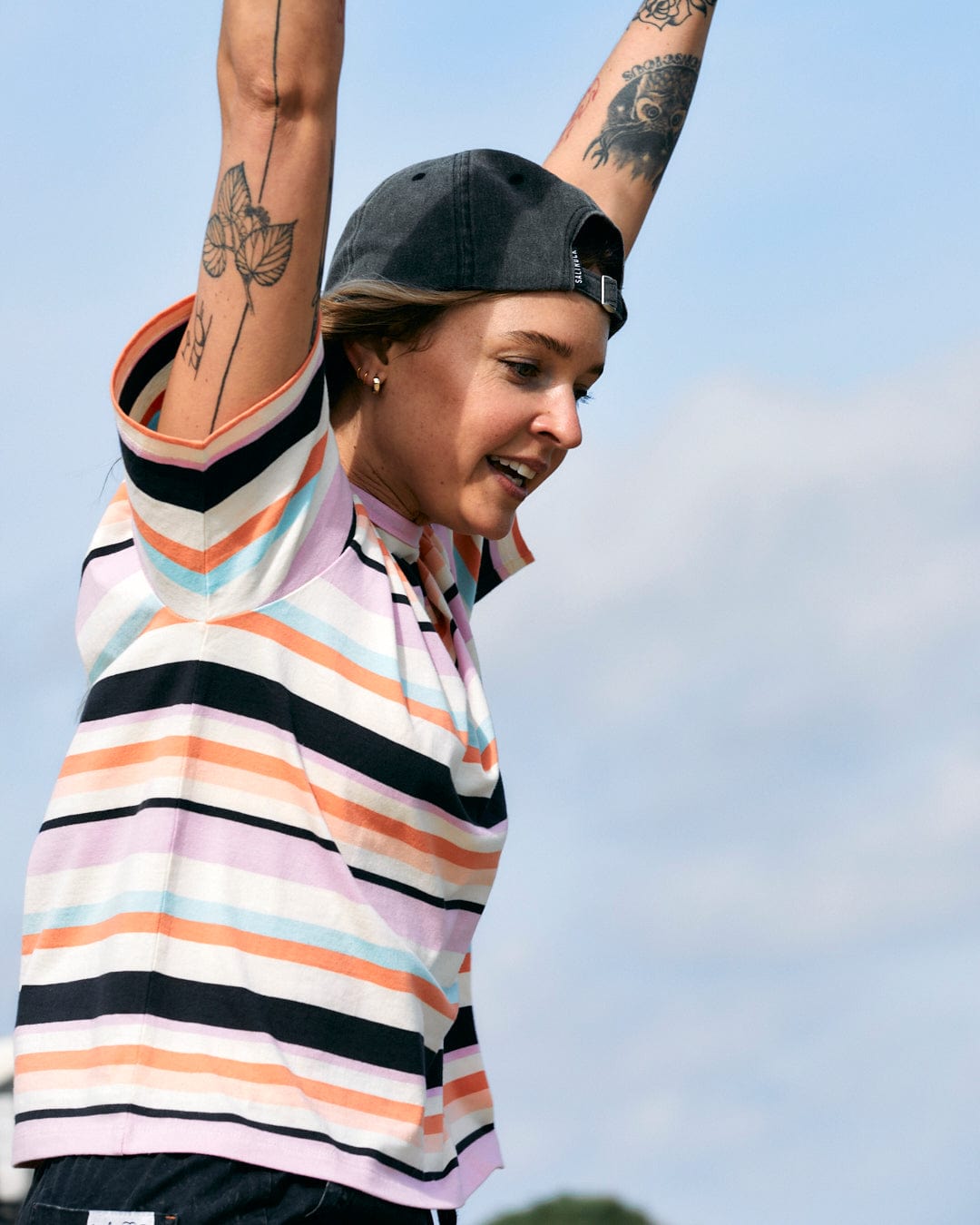 Woman with tattoos raising her arms in the air while wearing a Saltrock Juno - Womens Short Sleeve T-Shirt - Multi with embroidery and a cap.