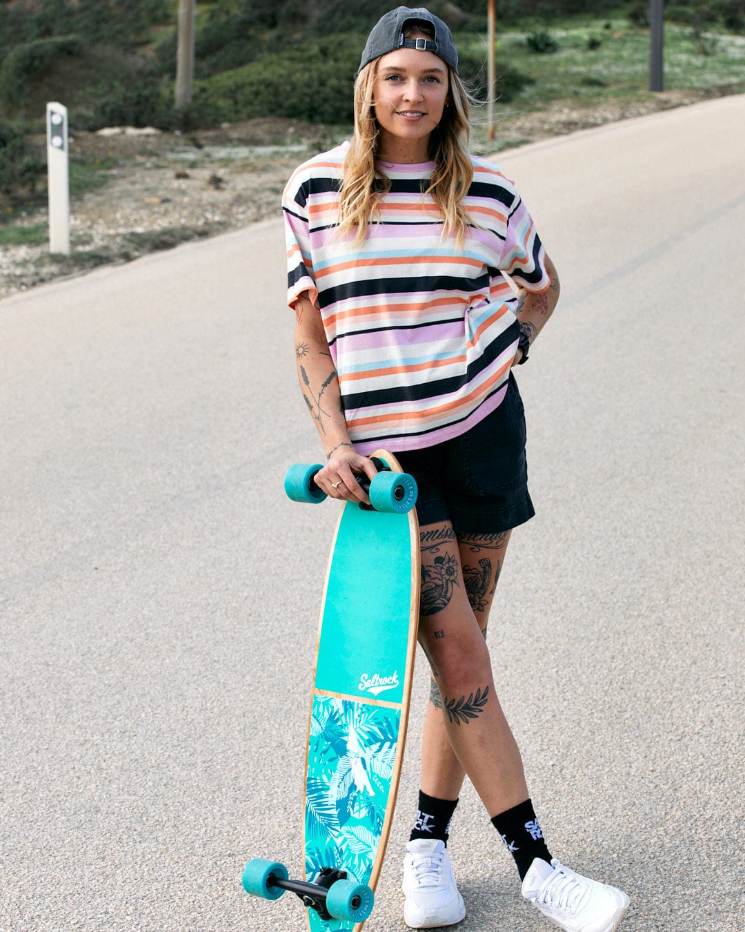A person standing with a skateboard on a path outdoors, wearing a Saltrock Juno - Womens Short Sleeve T-Shirt in Multi stripe pattern.