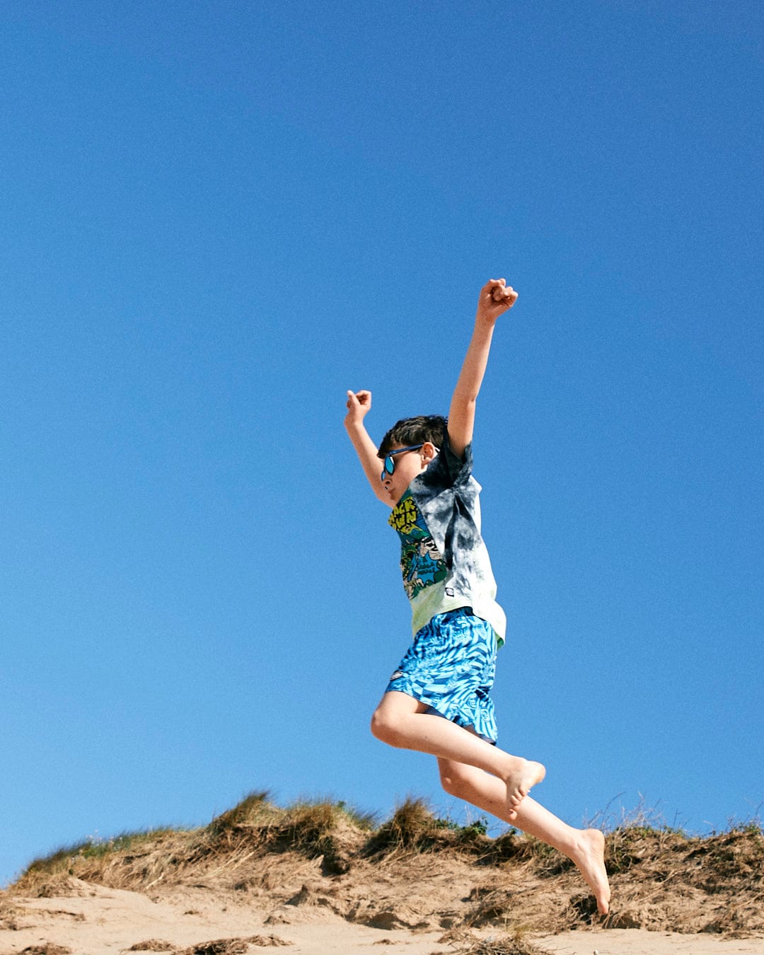A boy in blue shorts and a Saltrock Las Vegas Smackdown - Kids Glow in the Dark T-Shirt - Multi jumps energetically off a sandy hill against a clear blue sky.