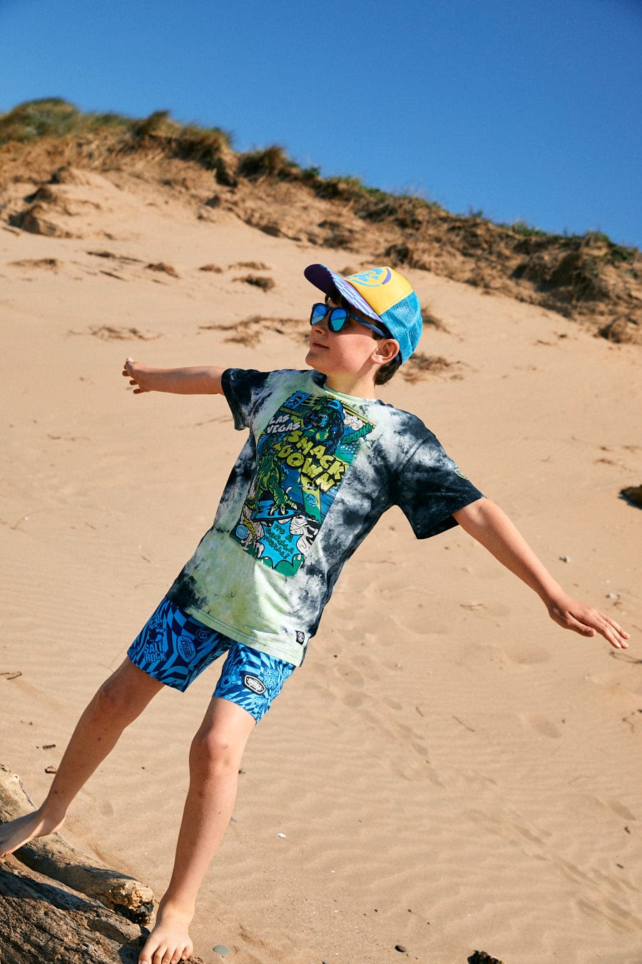 A child stands on a sandy beach wearing a cap, sunglasses, a Saltrock Las Vegas Smackdown - Kids Glow in the Dark T-Shirt - Multi, and tie dye pattern shorts, facing sideways with arms outstretched under a clear blue sky.