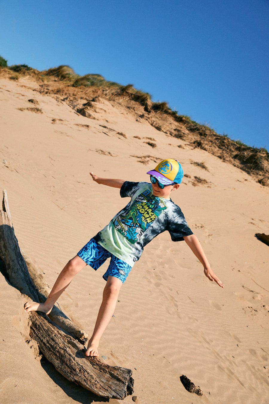 A child wearing a colorful cap, sunglasses, and a tie-dye shirt with glow in the dark graphics (Las Vegas Smackdown - Kids Glow in the Dark T-Shirt - Multi by Saltrock) balances on a log on a sandy beach with dunes in the background.