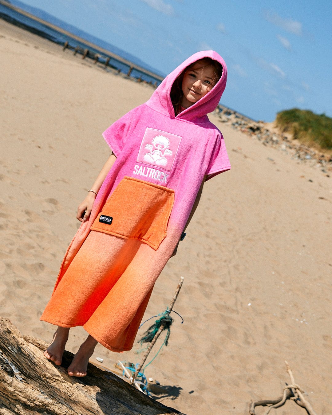A person wearing the Saltrock Tropic Dip - Kids Changing Towel - Pink/Orange stands on a sandy beach with scattered rocks. This ultra-absorbent, 100% cotton poncho is perfect for drying off quickly after a swim.