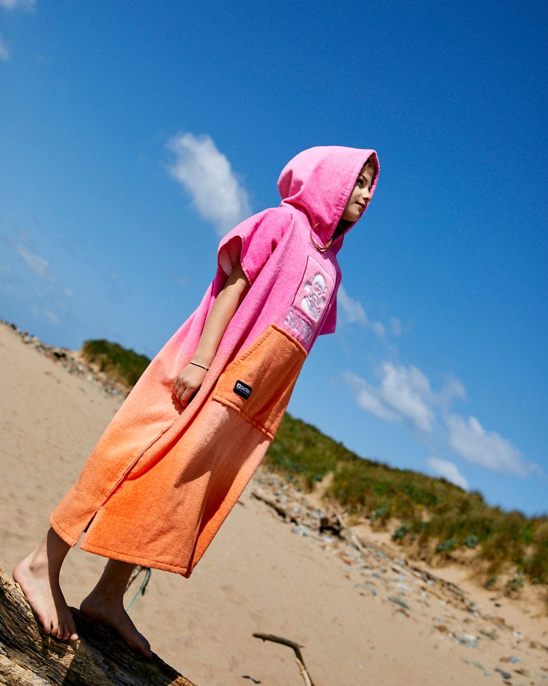 A child in a Saltrock Tropic Dip - Kids Changing Towel - Pink/Orange, made from ultra-absorbent 100% cotton, stands barefoot on a sandy beach with green grassy dunes and a blue sky with some clouds in the background.