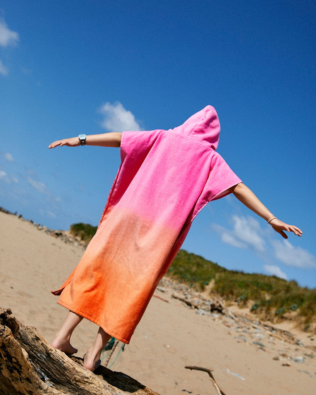 Person standing on a log at a beach, wearing a Saltrock Tropic Dip - Kids Changing Towel - Pink/Orange made of ultra absorbent 100% cotton, with arms outstretched under a clear blue sky.
