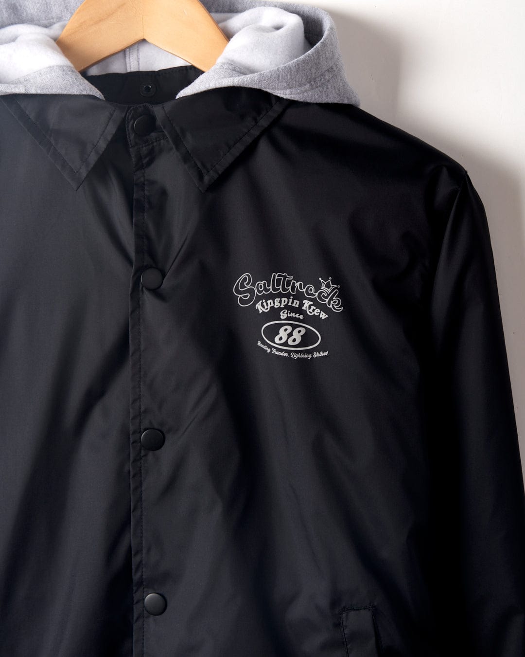 Close-up of a black, water-resistant Kingpin Krew Kids Coach Jacket with a gray hoodie and a logo on the left side reading "culture cafe australia 88 sydney diner.