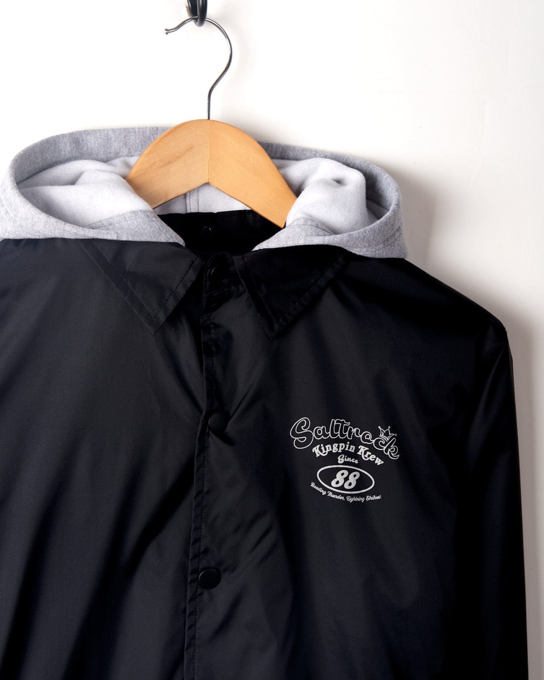 Black and gray water-resistant Kingpin Krew - Kids Coach Jacket with embroidered logo "sailor's creek 88" hanging on a wooden hanger against a white wall. (Saltrock)
