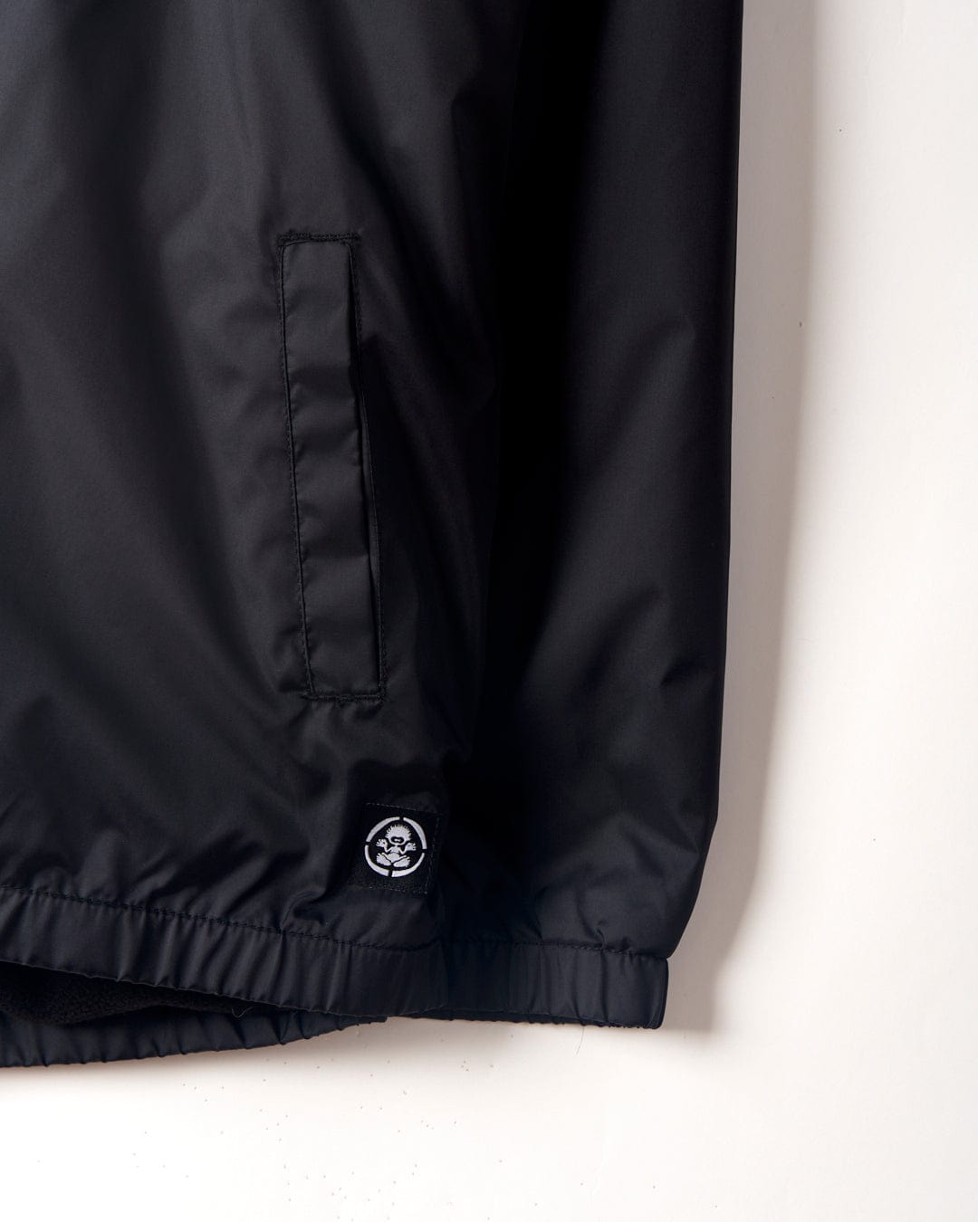 Close-up of a Saltrock Kingpin Krew - Kids Coach Jacket - Black hem with an embroidered logo patch on the lower right side and a vertical stitched pocket detail on textured fabric.