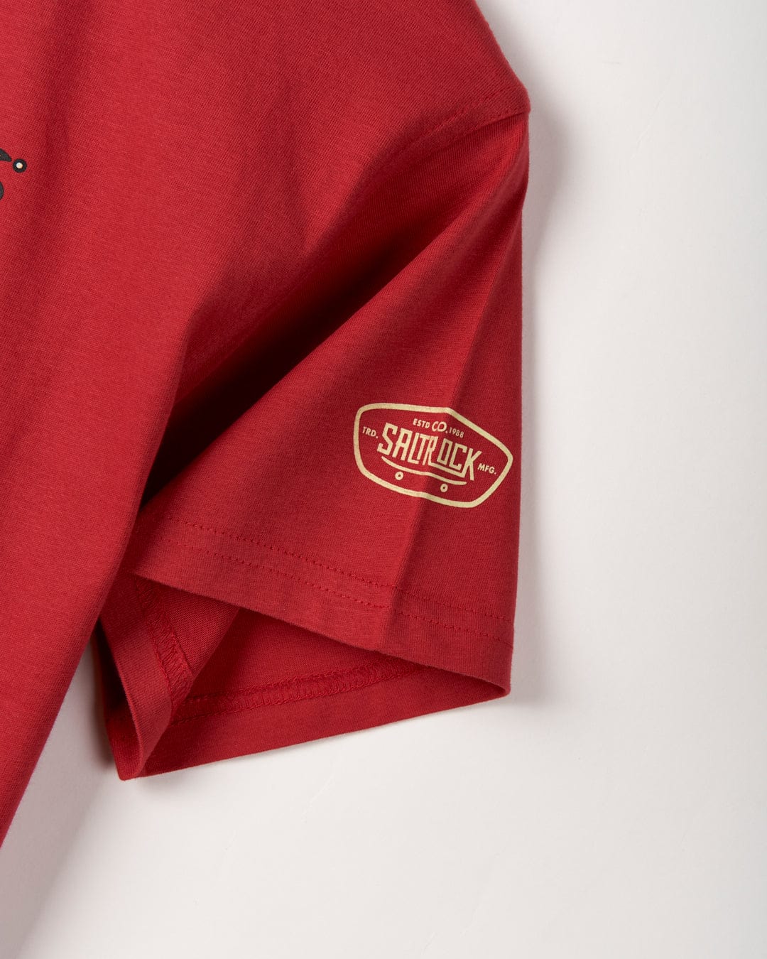 Close-up of a red shirt sleeve with a small oval logo that reads "Saltrock" in white text on a gold background, featuring a relaxed fit design for ultimate comfort. This is the Kingpin Krew - Kids Short Sleeve Glow in the Dark T-Shirt.