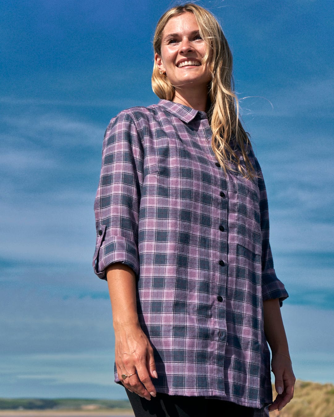 A woman wearing a Saltrock Kizzie Womens Check Boyfriend Shirt in Purple standing on a beach.