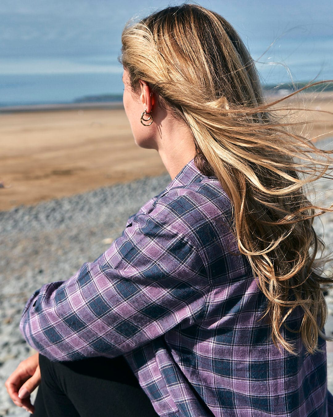 A woman sitting on the beach in a relaxed fit, Saltrock Kizzie - Womens Check Boyfriend Shirt - Purple, with her hair blowing in the wind.