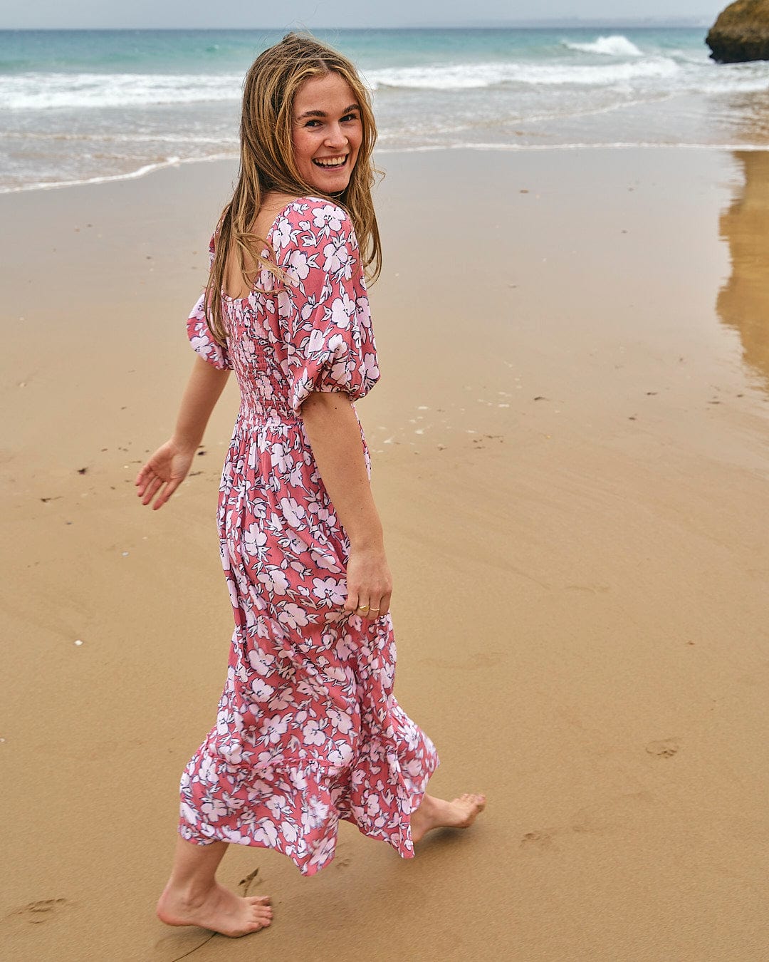 A woman walking on the beach in a Saltrock Larran - Womens Shirred Dress - Mid Pink.