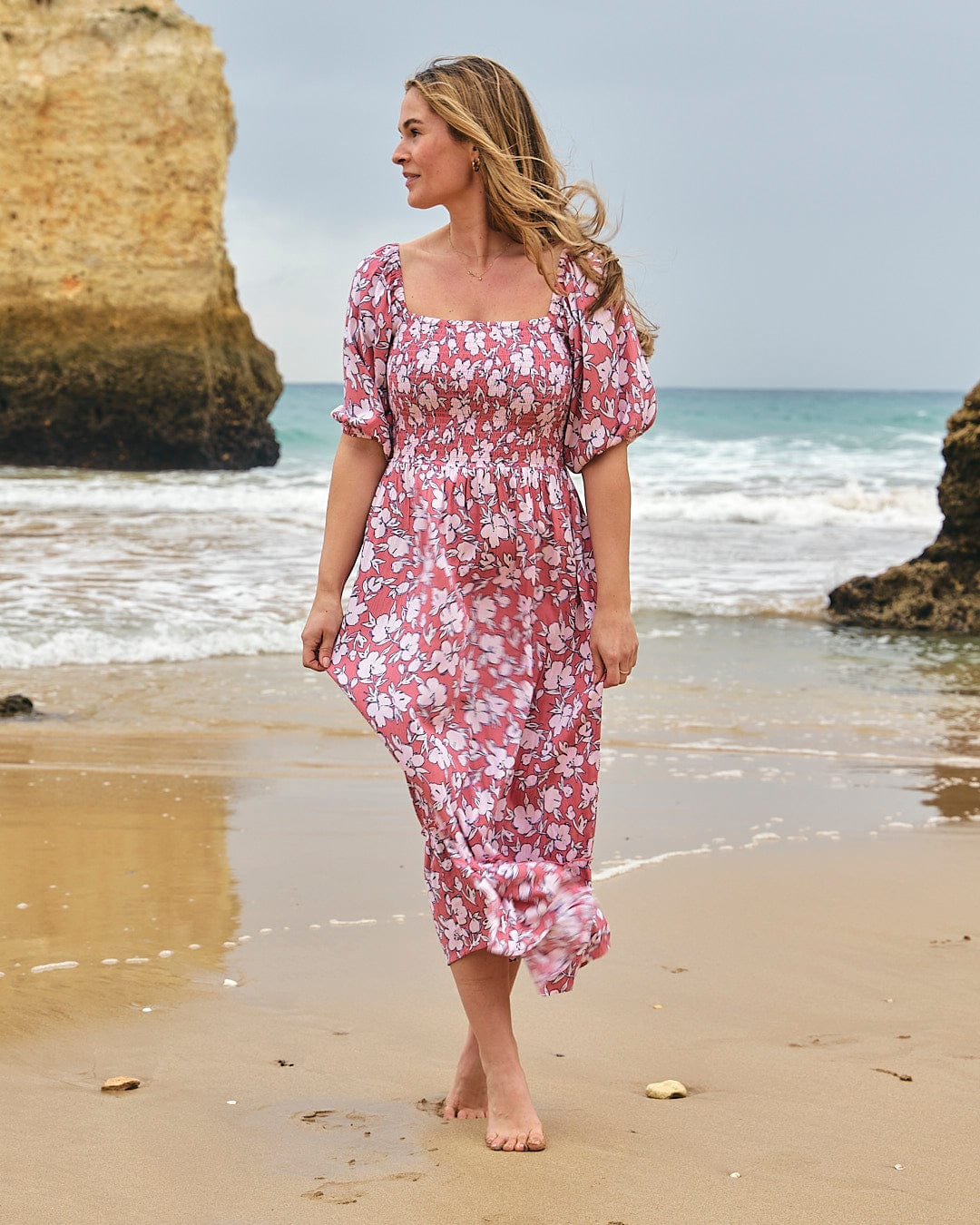 A woman walking on the beach in a Saltrock Larran - Womens Shirred Dress - Mid Pink.