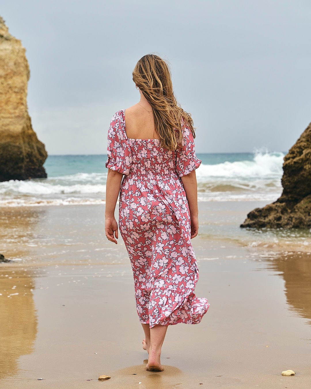 A woman walking on the beach in a Saltrock Larran - Womens Shirred Dress - Mid Pink.