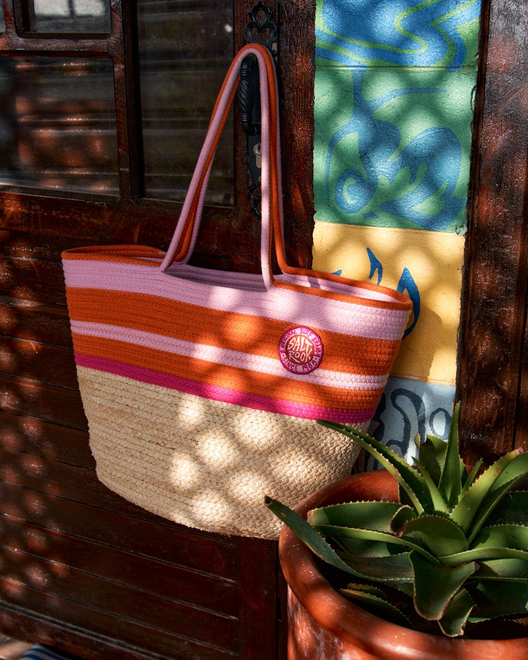 Colorful retro Las Cora Straw Beach Bag - Cream hanging on a wooden door next to a succulent plant and a brightly patterned cloth.