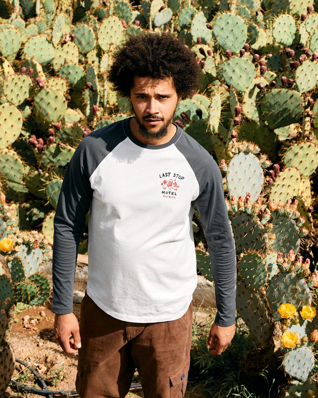 A man with curly hair stands in front of cacti, wearing a gray Saltrock Last Stop long sleeve raglan t-shirt and brown shorts.