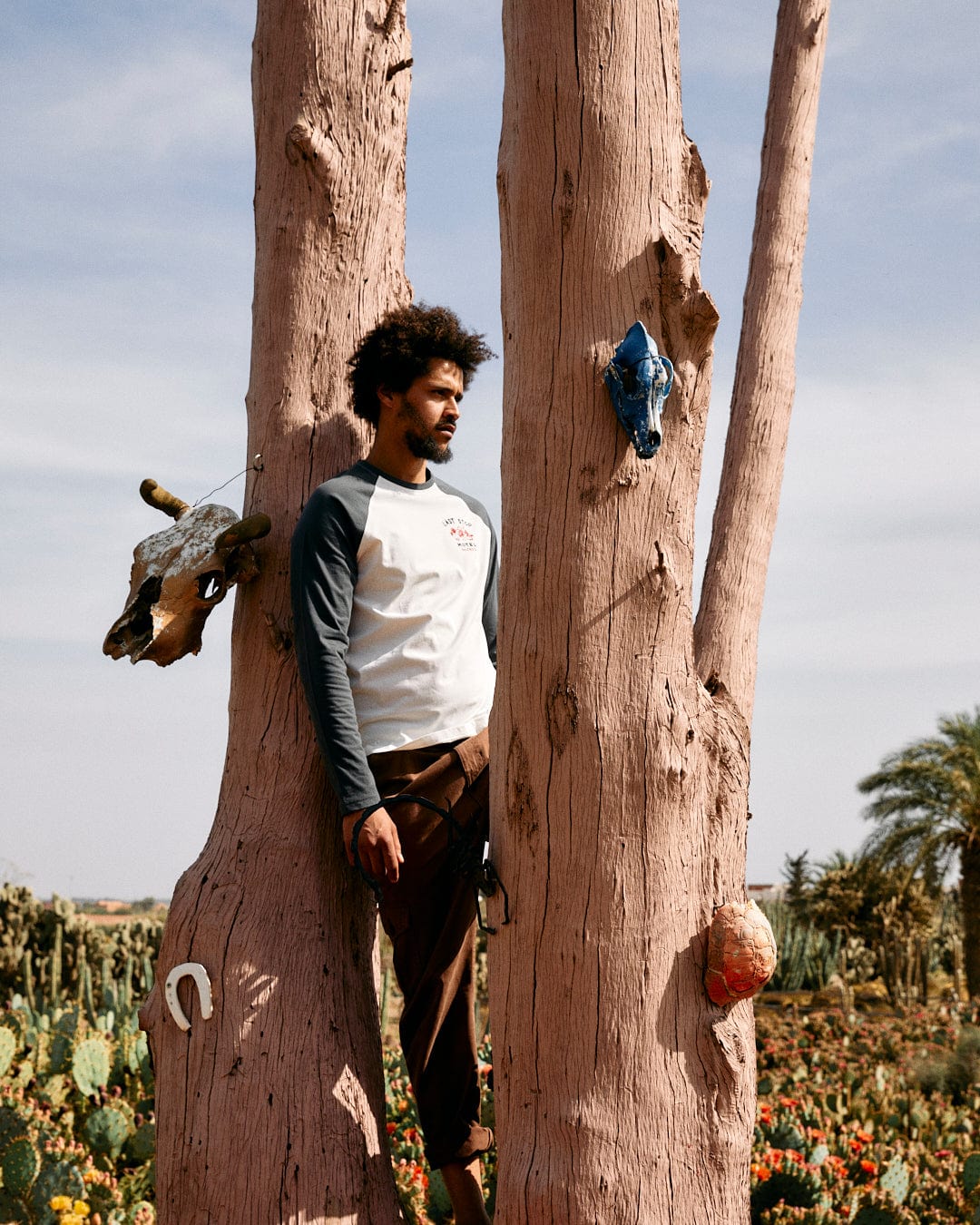 A man with curly hair stands between two rugged trees decorated with various objects, in a sunlit desert garden, wearing a Last Stop - Long Sleeve Raglan - White by Saltrock.