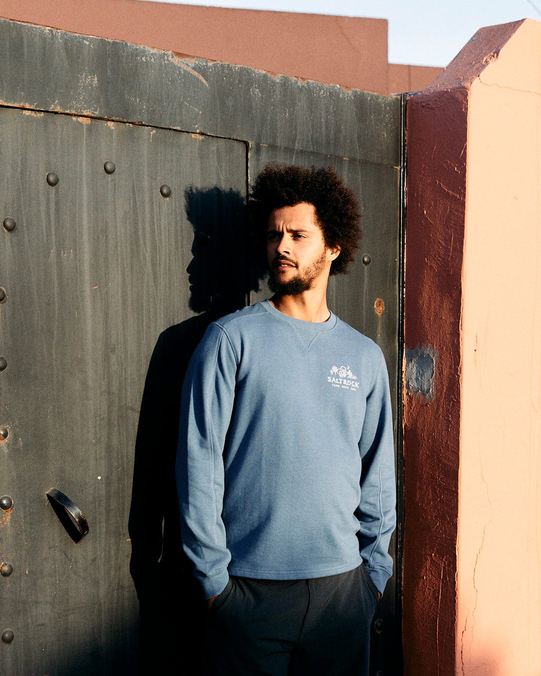 A man with an afro stands beside a rustic metal door at the Last Stop Motel, wearing a Saltrock blue long-sleeve shirt with a crew neckline, gazing to the side in soft sunlight.