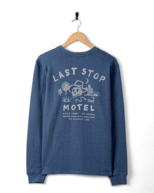 Blue Last Stop Motel mens sweatshirt made of peached soft material with "Last Stop Motel" skull graphic design, hanging on a white wall.