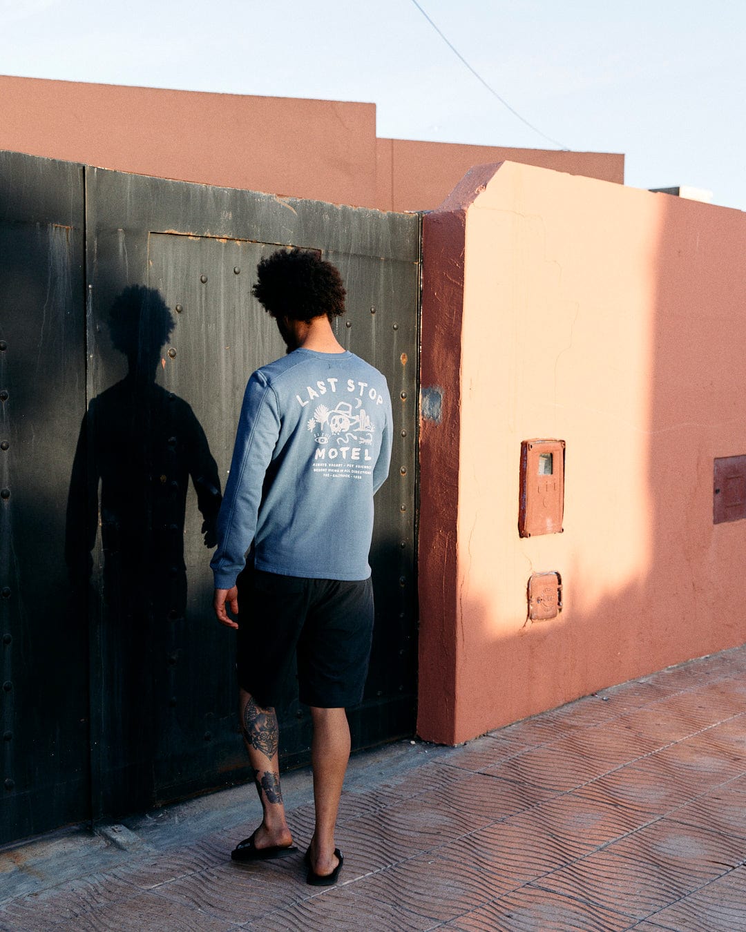 A person with curly hair wears a Saltrock Last Stop Motel - Recycled Mens Sweatshirt in Blue and stands next to a rustic green door on a peach-colored wall, casting a shadow.