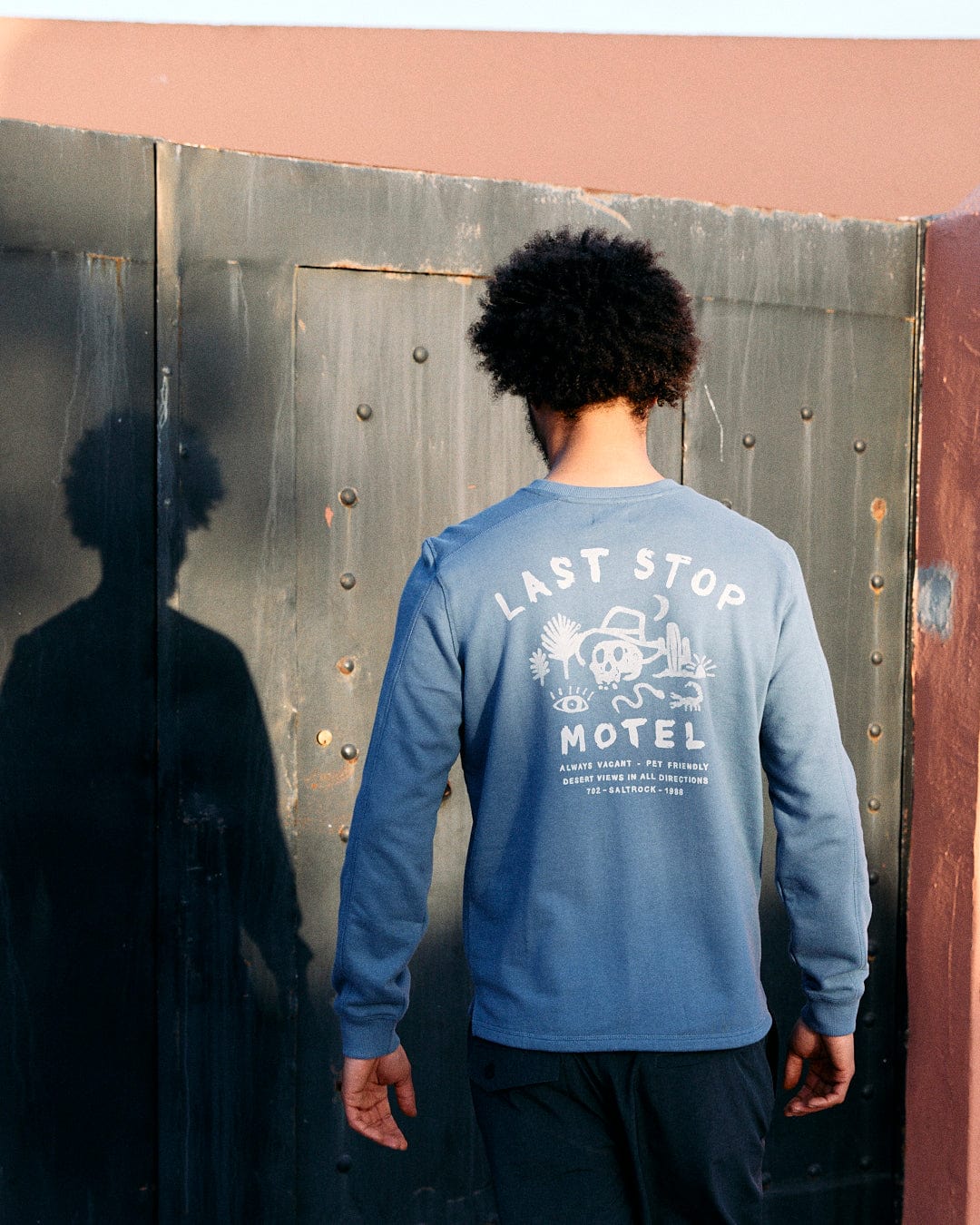 A person with an afro standing with their back to the camera, wearing a Saltrock blue sweatshirt with "Last Stop Motel" print and a crew neckline, in front of a metal door.