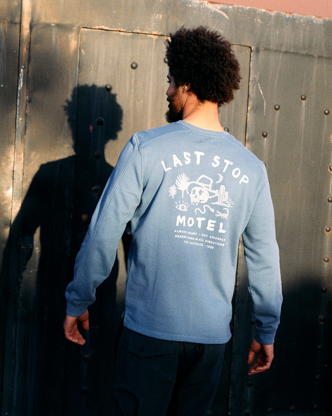 A man with an afro facing away from the camera, wearing a blue Saltrock sweatshirt made of peached soft material with "Last Stop Motel" graphic, standing by a metal wall.