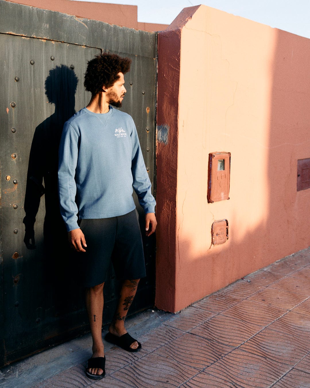 Man with curly hair standing beside a pink wall and green door during sunset, wearing a Last Stop Motel - Recycled Mens Sweatshirt in Blue from Saltrock, black shorts, and flip-flops.