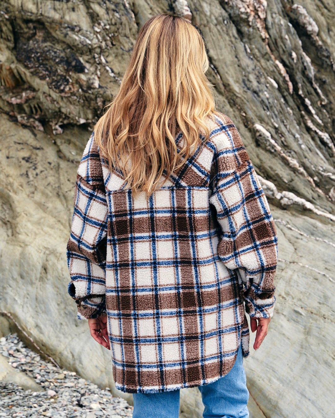A woman wearing jeans and a Saltrock Laurie - Womens Check Sherpa Fleece Coat - Cream standing on a rocky beach in cold weather.