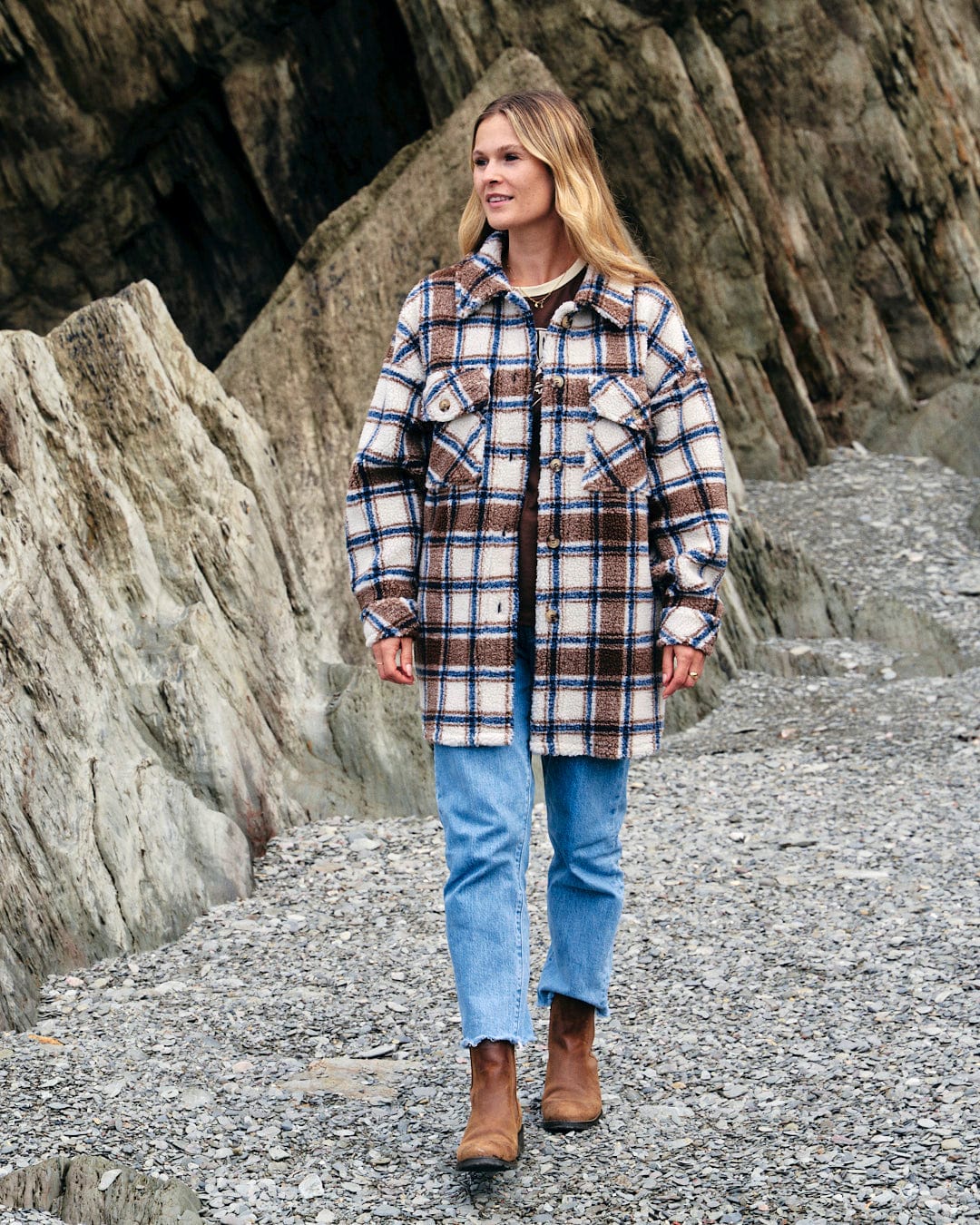 A woman wearing a Laurie - Womens Check Sherpa Fleece Coat - Cream by Saltrock walking on rocks in cold weather.