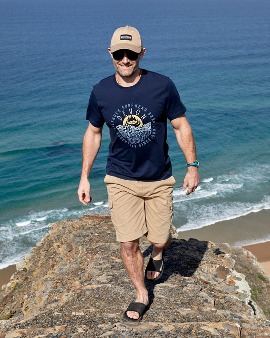 A man in a Saltrock Layers Devon - Mens Short Sleeve T-Shirt - Blue hat and t-shirt, made from peached soft material, standing on top of a cliff overlooking the ocean.