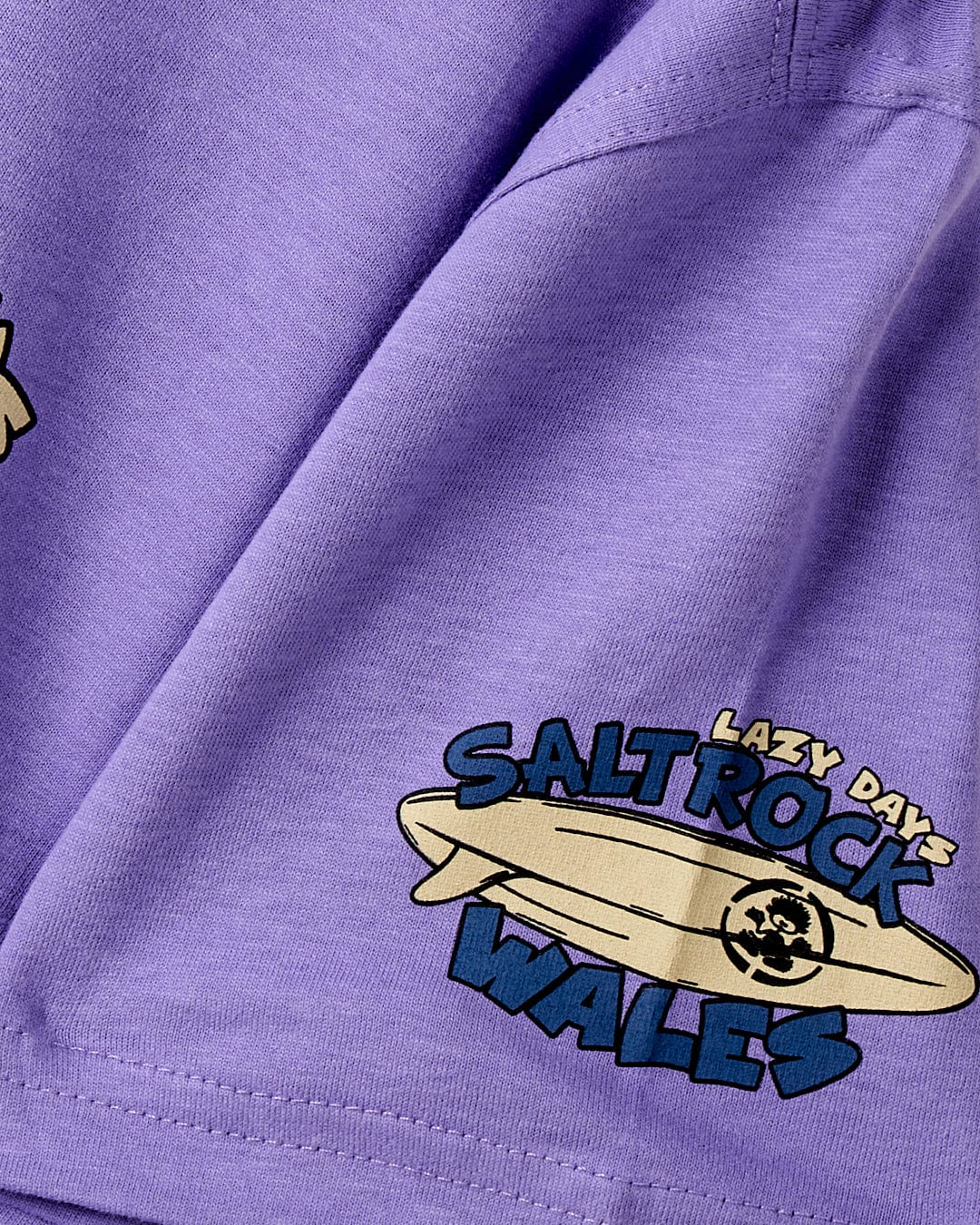 Close-up of a purple cotton material with a "Saltrock" logo patch featuring surfing graphics on the Lazy Location Wales - Kids T-Shirt - Purple.