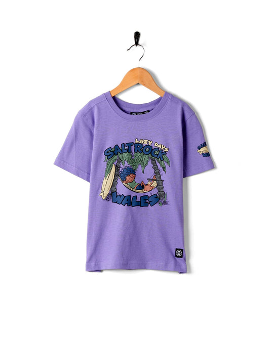 Saltrock Lazy Location Wales Kids T-Shirt in Purple with surfing graphics print hanging on a white wall.