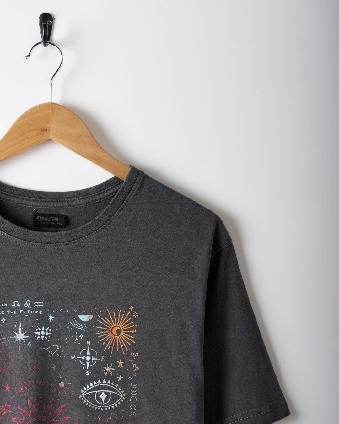 A washed grey, oversized T-shirt by Saltrock, featuring a mystical gradient print of colorful celestial symbols, hangs on a wooden hanger against a plain white wall.