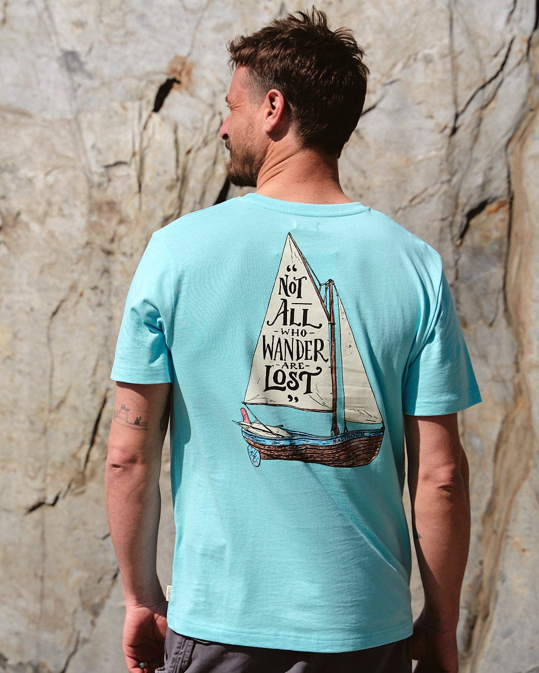 A man wearing a Lost Ships - Mens Short Sleeve T-Shirt in Turquoise by Saltrock, with a sailboat on it made of cotton.