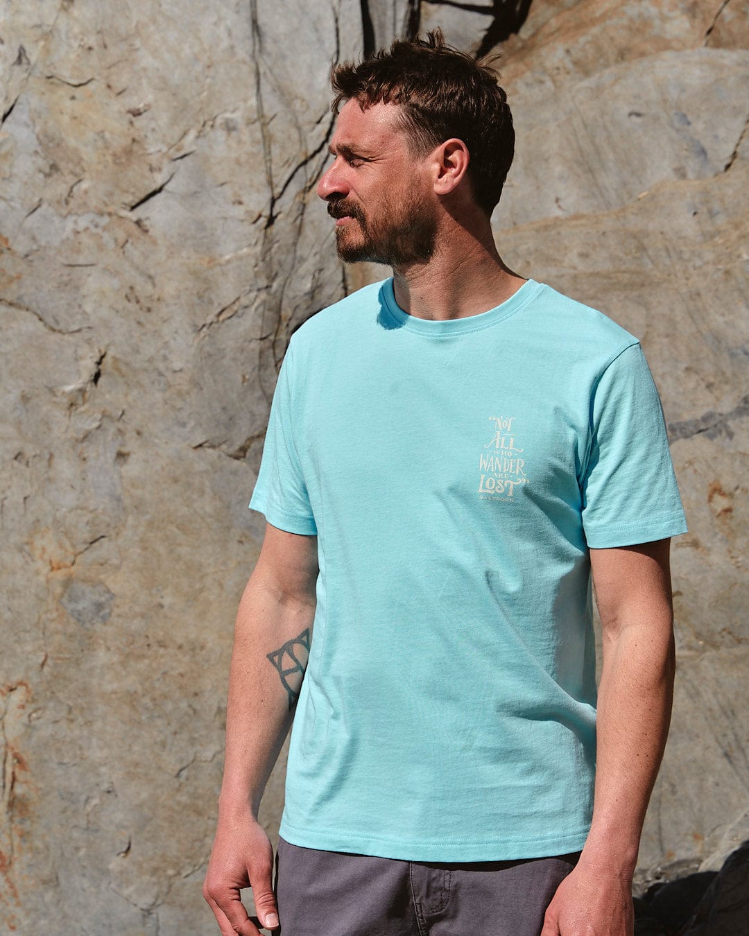 A man in a Lost Ships - Mens Short Sleeve T-Shirt in Turquoise by Saltrock stands in front of a rock.