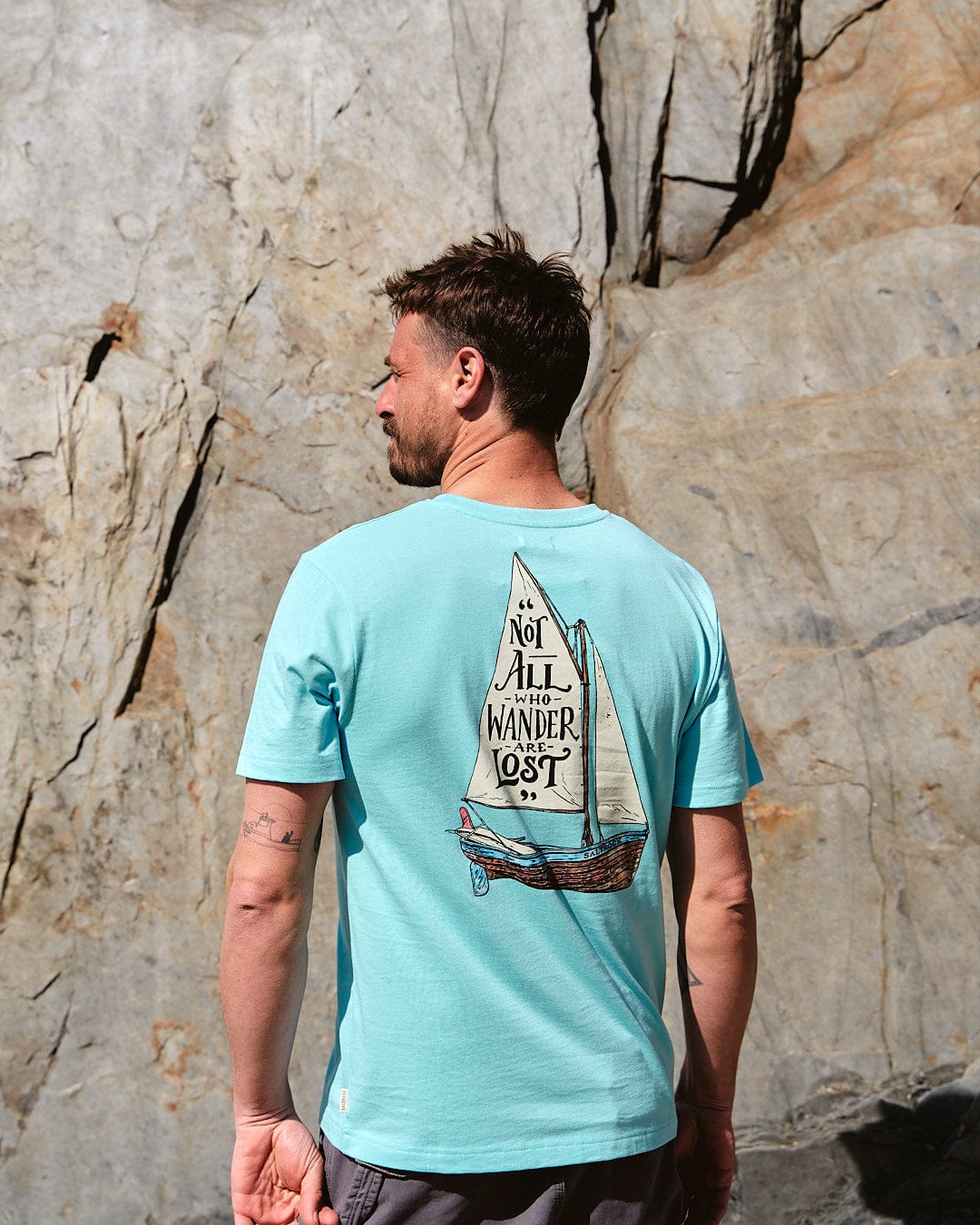 A man wearing a Lost Ships - Mens Short Sleeve T-Shirt in Turquoise by Saltrock with a sailboat on it.