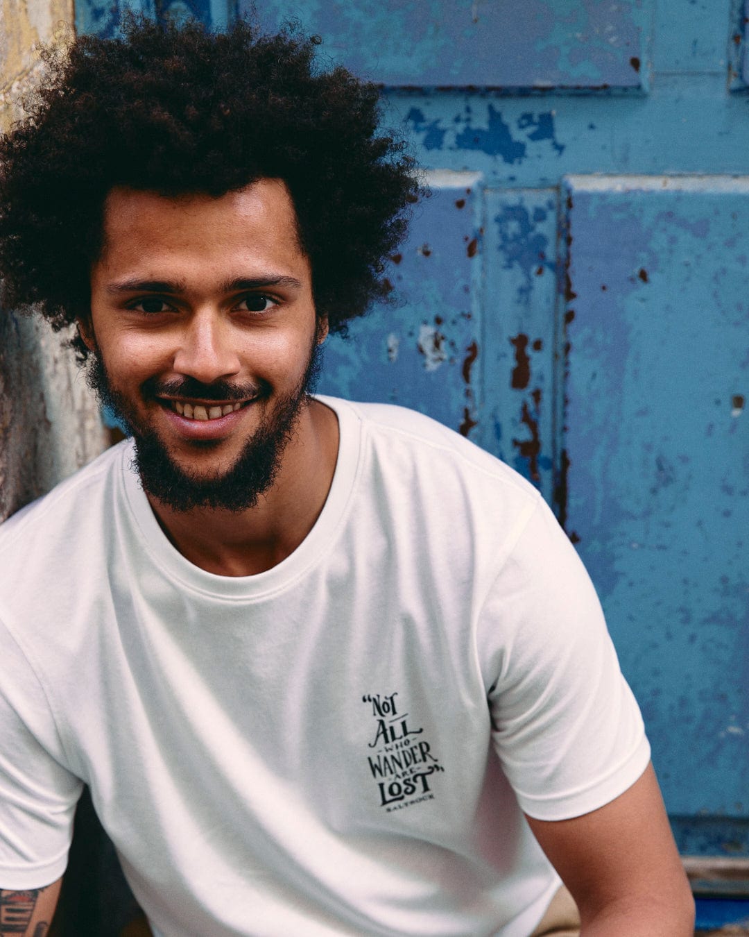 A man with curly hair smiles, wearing a white Saltrock Lost Ships Short Sleeve T-Shirt with a crew neckline against a blue rustic door background.