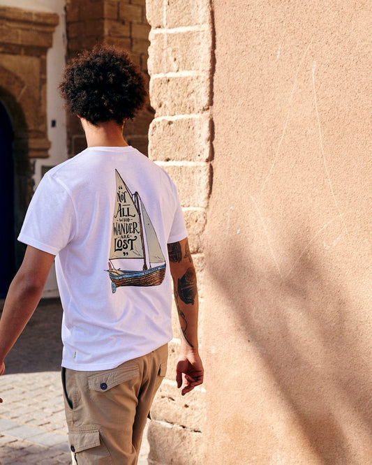 A man with curly hair walks past a sandy-colored wall, wearing a Saltrock white t-shirt with a sailboat design and the text "not all who wander are lost," featuring a crew neckline.
