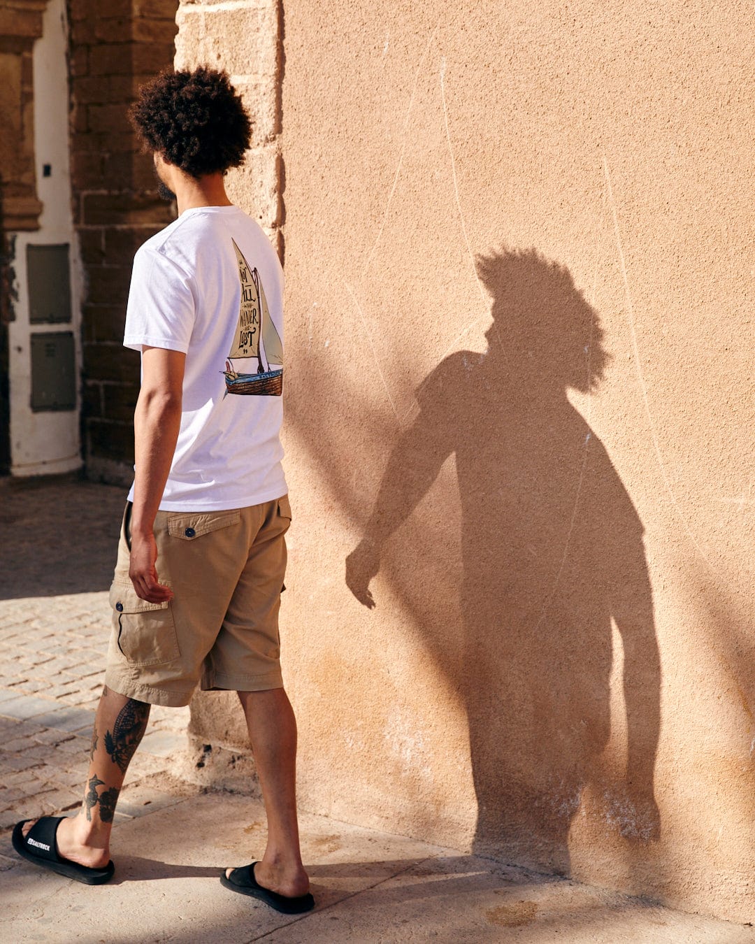 A young man with curly hair looks at his shadow on a sunlit beige wall, wearing a Saltrock Lost Ships Short Sleeve T-Shirt in White, khaki shorts, and sandals.