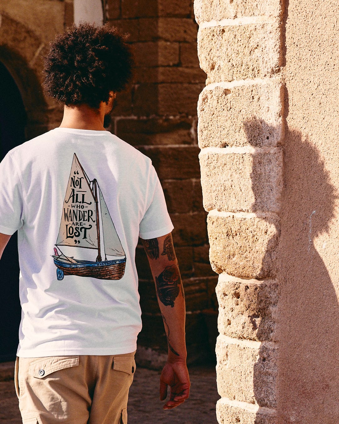 A man with curly hair and a tattooed arm wearing a crew neck t-shirt with a Saltrock Lost Ships Short Sleeve T-Shirt in White design, walking by a stone wall, holding sunglasses.