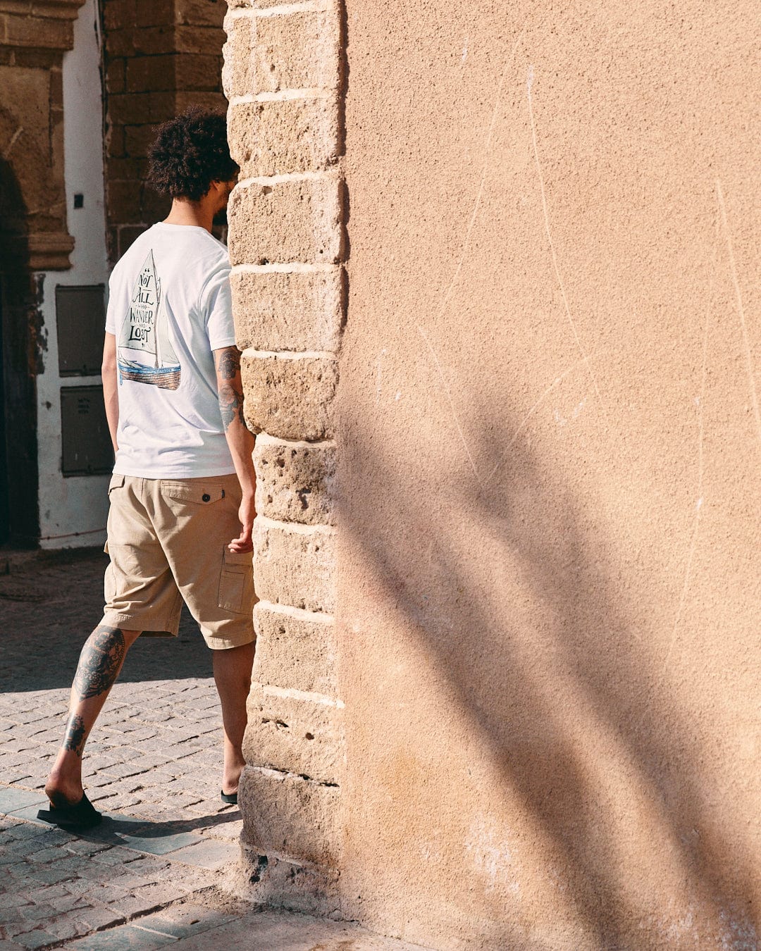 A man with curly hair and tattoos walks past a textured stone wall on a sunny day, casting a shadow. He's wearing a white Saltrock Lost Ships Short Sleeve T-Shirt with a crew neckline.